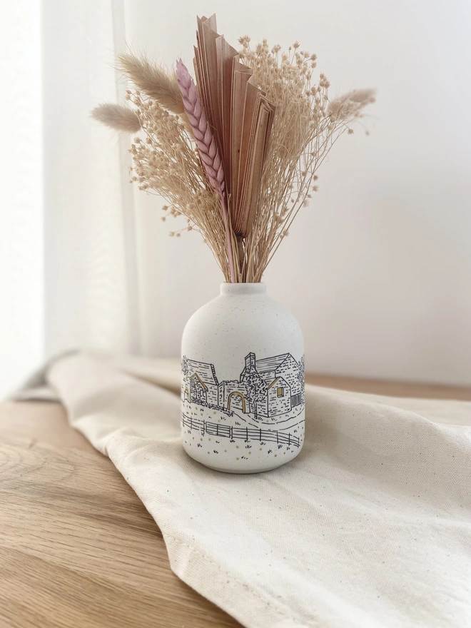 A ceramic vase featuring a black line drawing of a wedding venue with a dried flower bouquet