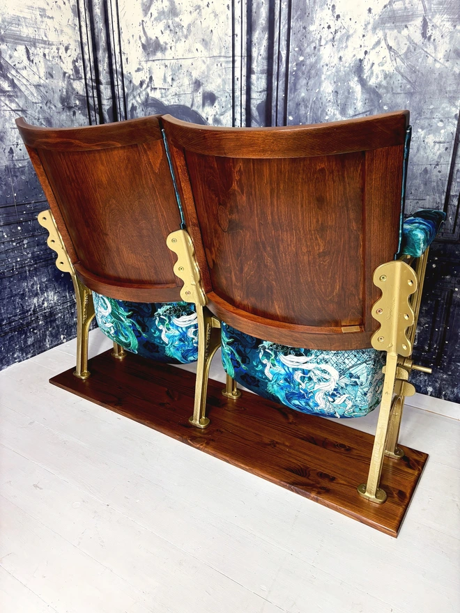 Wood backs on a set of vintage cinema seats, upholstered in blue mermaid velvet.  Seats are on a wooden plinth sat on a white wooden floor with a blue marbled walls