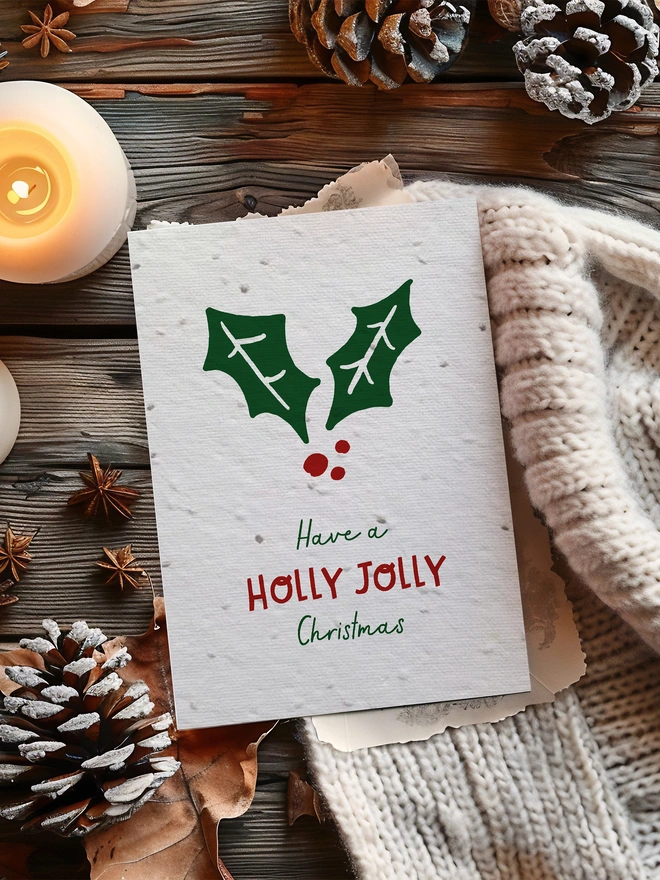 A Christmas card with an illustrated holly leaf with ‘Have a Holly Jolly Christmas’ underneath. The card is placed on a wood background with a lit candle, pine cones and cosy blanket.