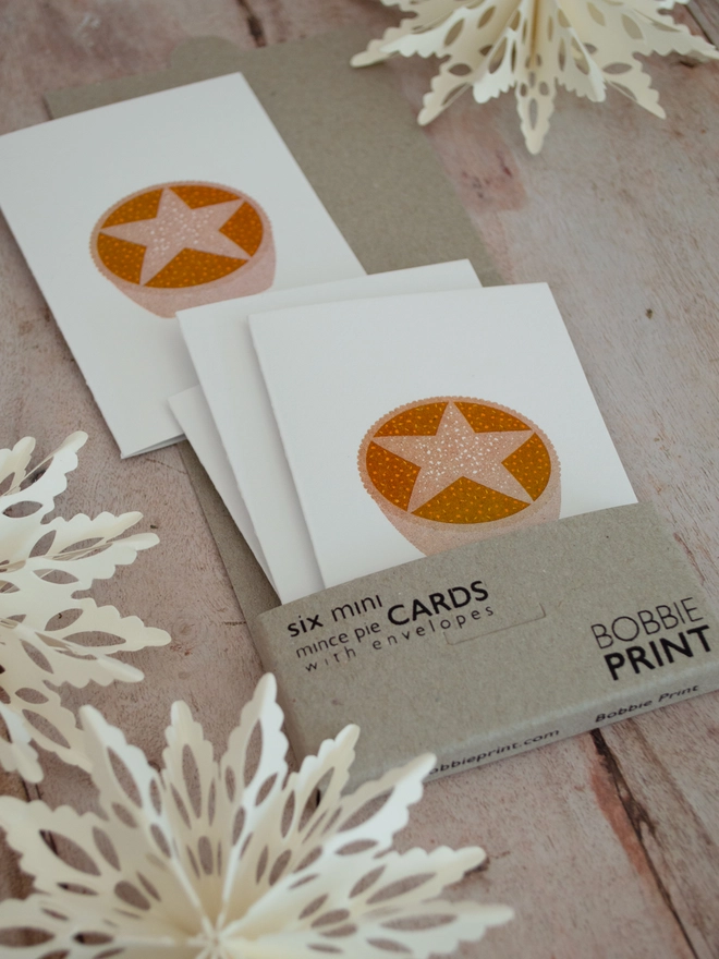A boxed pack of Six Mini Mince Pie Christmas Cards lies open on a festive wooden table, several small white cards spill out, each one risograph printed with a cute little mince pie illustration. aper cut snowflakes lie around the box and cards.