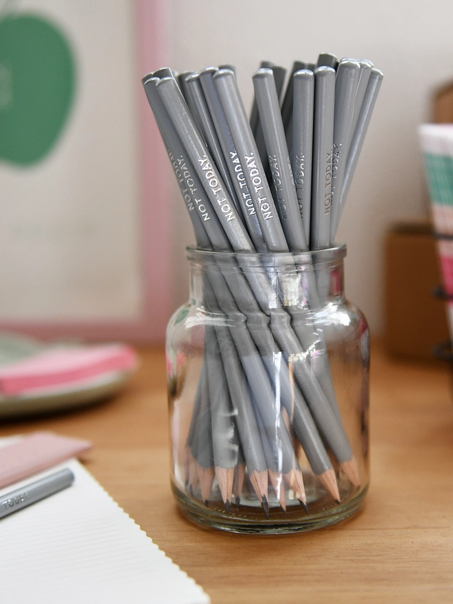 A glass jar full of grey pencils stands on a wooden desk surrounded by various stationery items.