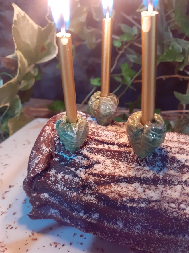 Close-up of three green ceramic sprout cake candle holders with glowing gold candles, displayed on a chocolate yule log dusted with icing sugar.