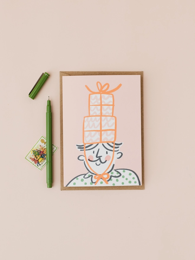 Images shows a Christmas card which features an illustration of a man wearing a gift shaped party hat on a pink background.
