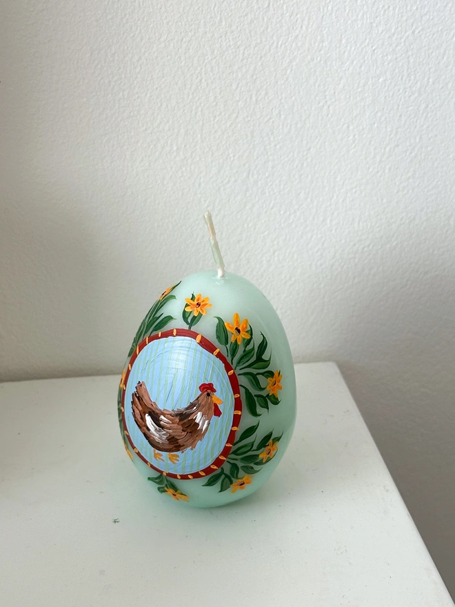 Hand Painted Easter Egg Candle Hen & Flowers, an Easter egg candle sitting on a plain surface. It's Easter and floral design wraps around the egg. 