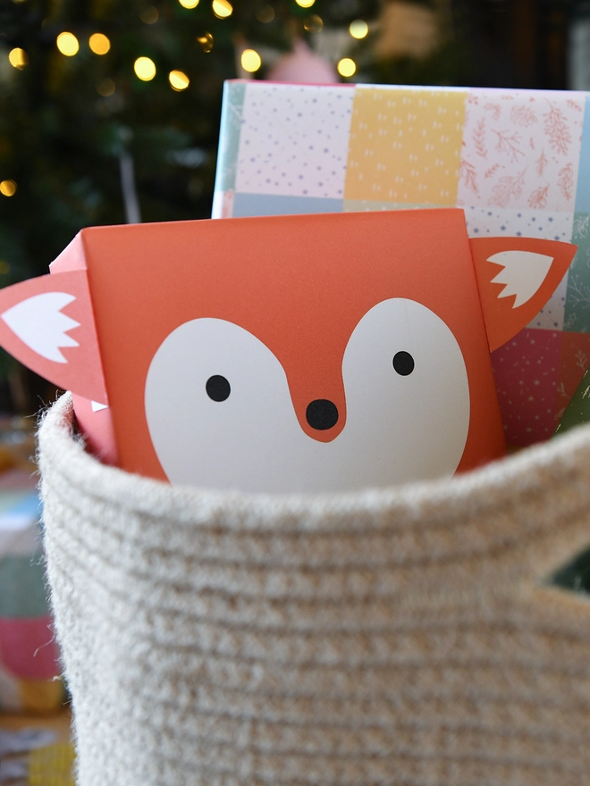 A gift wrapped as an orange fox is tucked into woven basket beside other wrapped gifts in front of a Christmas Tree.