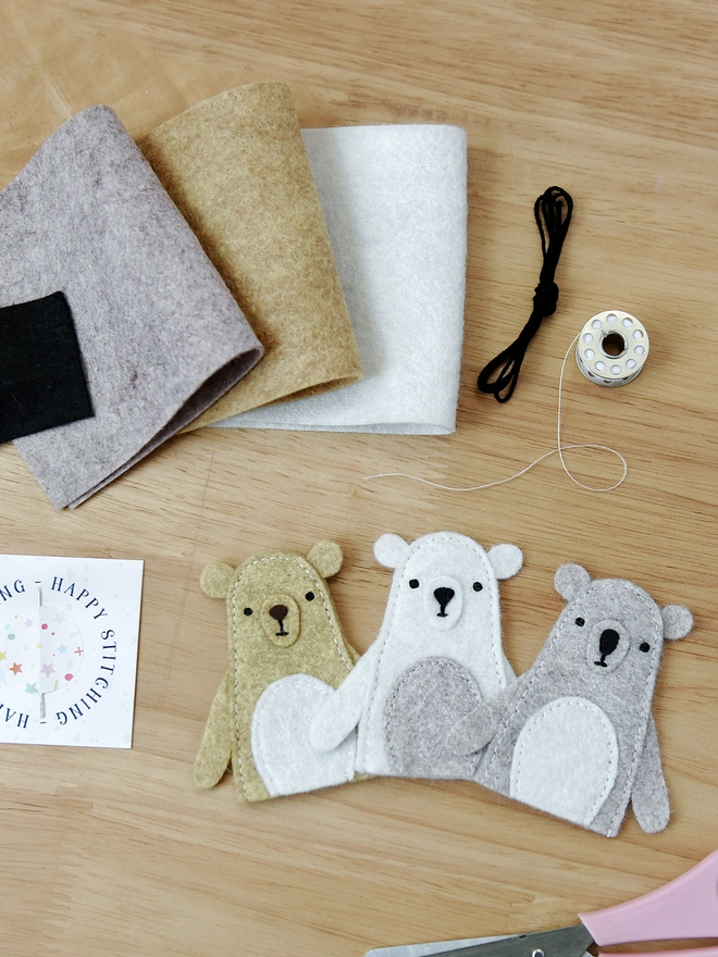 Three handmade felt bear finger puppets lay on a wooden desk with various craft kit materials beside them.