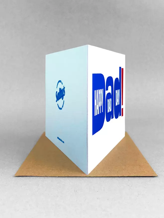Studio shot of white landscape greetings card on light grey background, rear view showing the folded spine of the card towards us, the front of the card to the right and the rear in view with Salty’s logo and bumf. The design is typography of the word Dad! With happy Dad Day written within the letters. The writing is printed in royal blue with an oversized exclamation mark in red to the right. The card is stood on a beige kraft envelope.