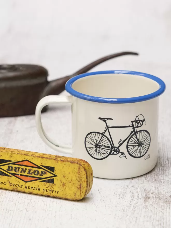 Picture of a Cream Enamel Mug with a Blue Rim with a Bicycle design etched onto it, taken from an original Lino Print