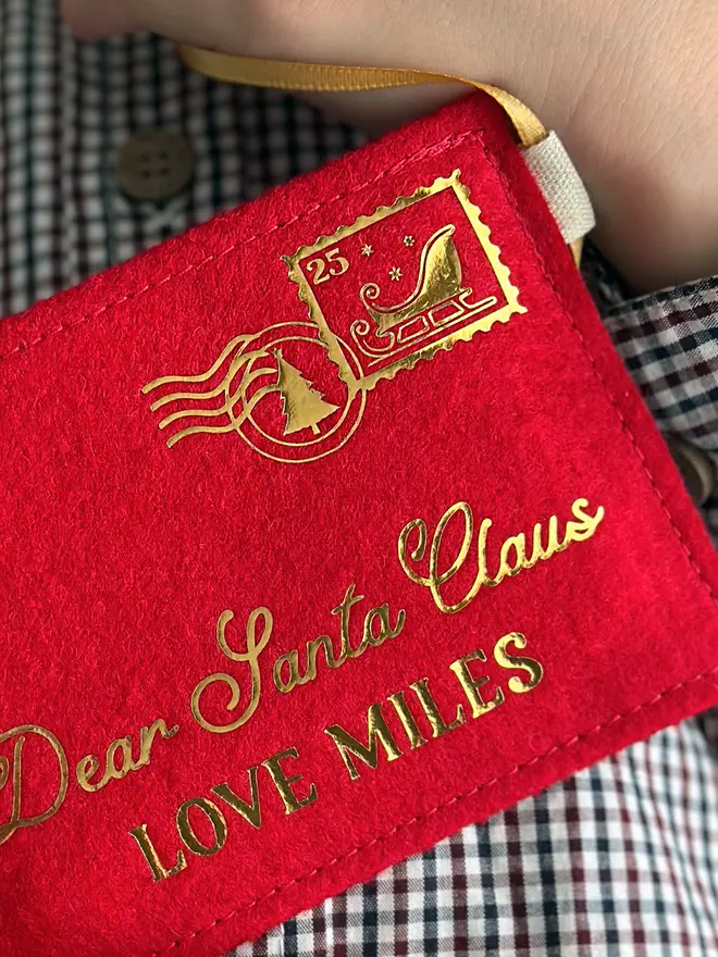 A child wearing a gingham shirt is holding a handmade red felt Christmas envelope that reads "Dear Santa Claus, Love Miles" in gold writing with a golden sleigh stamp in the top right hand corner.