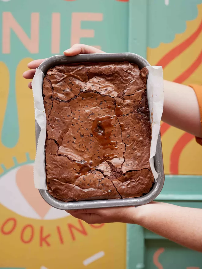 Freshly baked sea salted caramel brownies in a baking tray being held in front of a colourful background