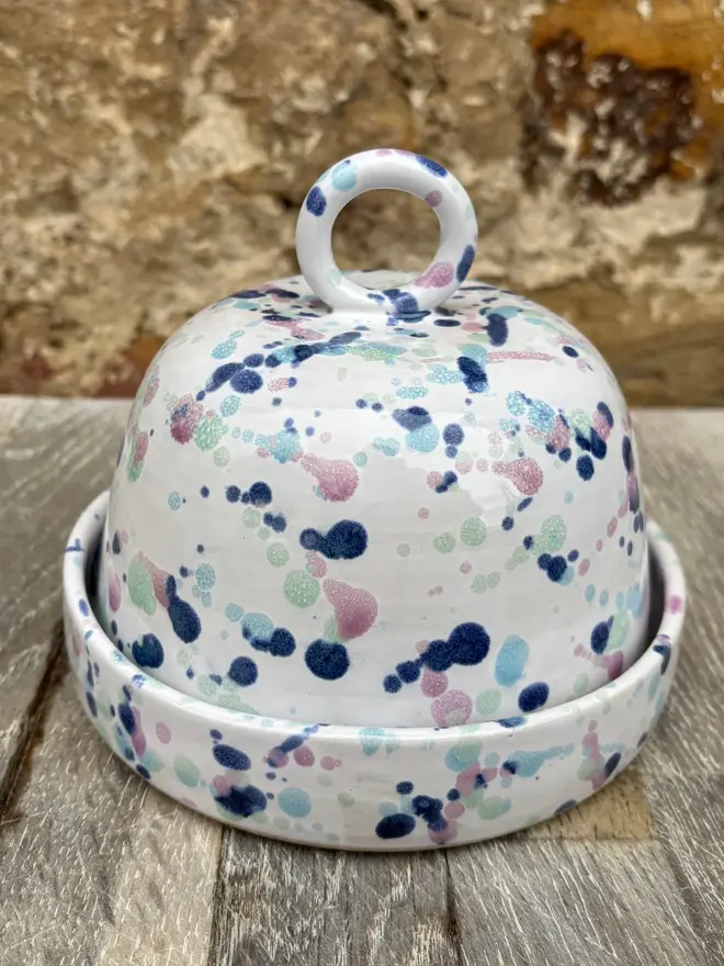 Sweetpea Butter Dish, a colourful butter dish sitting on a wooden surface against a brick backdrop. 