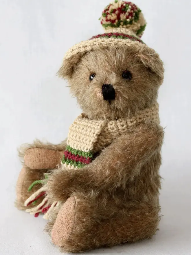 Brown teddy bear sitting upright against a white background, wearing a knitted beige hat and scarf with green and burgundy details.