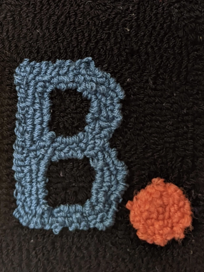 close up of a knitted upper case 'B' in blue wool with an orange full stop on a black background