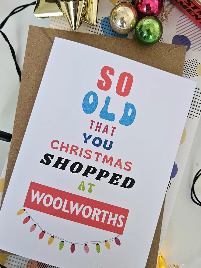 A photograph of the front of a humorous A6 greeting card illustrated using brightly coloured text and the retro shop logo. The text says so old you Christmas shopped at Woolworths with string lights