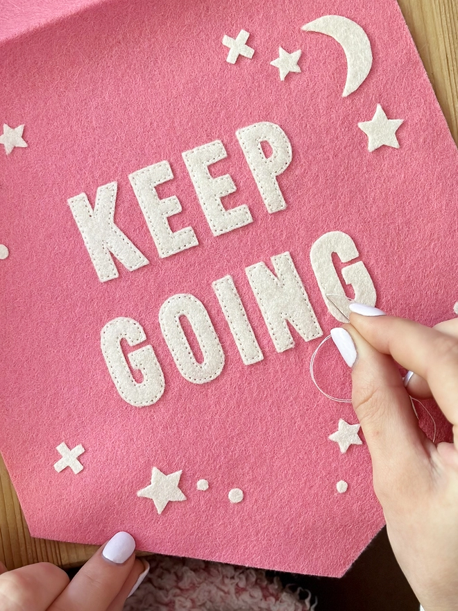 A pink felt banner flag is being sewn over a wooden desk. White felt stars are scattered around the words Keep Going in the centre of the flag.
