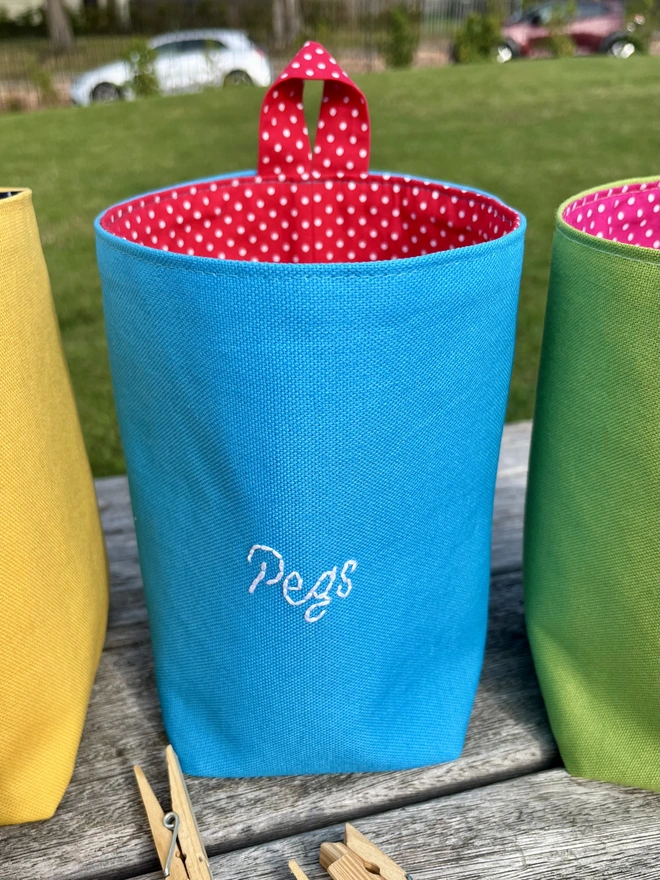 Large blue cotton clothes peg bag with red polka dot lining and hanging loop standing on a garden bench.