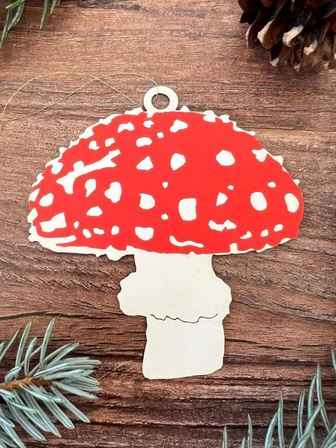 A red and white paper mushroom decoration, threaded with a thin gold thread loop is resting on a dark wood surface surrounded by spruce and pinecones.
