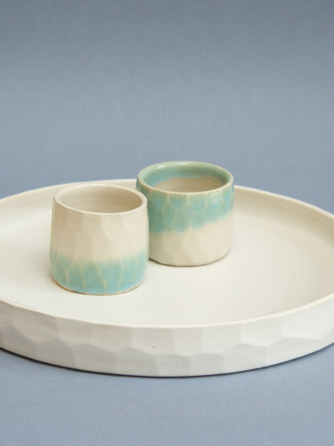 Two straight sided ceramic cups next to eachother on a ceramic matt white low, shallow tray.  One has a turquoise top half fading into a glossy cream bottom half.  The other cup as the opposite, with the top half being glossy cream and the bottom half being turquoise.