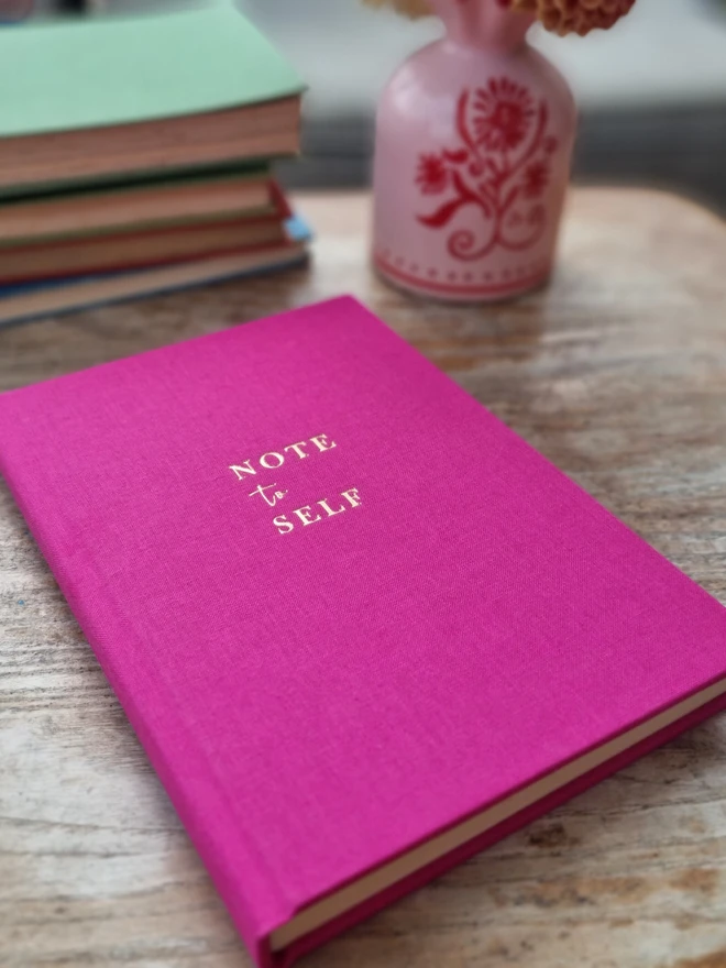 A bright pink notebook printed with the words 'Note to Self' in gold lettering rests on a wooden table top.
