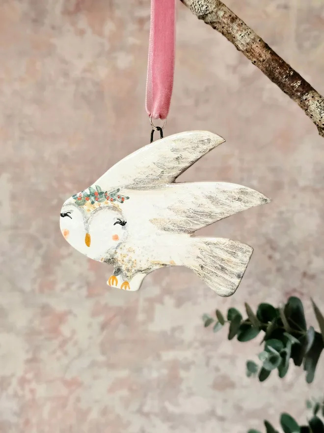 Decorative owl ornament with floral crown hanging from a branch on a soft pink background.