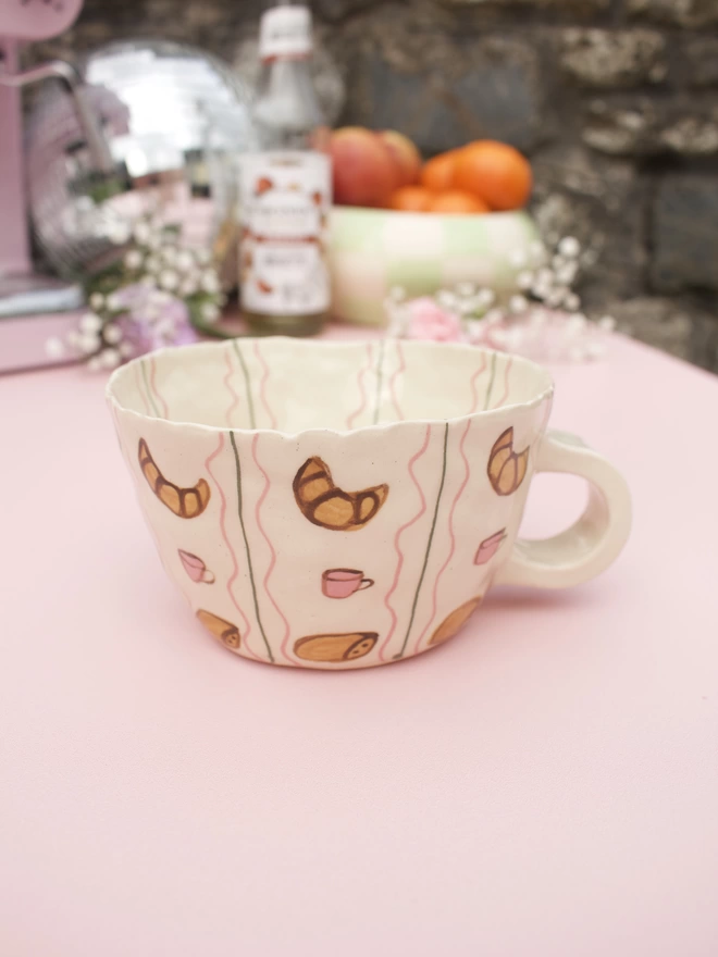handmade stoneware mug decorated with handpainted croissants, coffee cups and pain au chocolat in a striped pattern