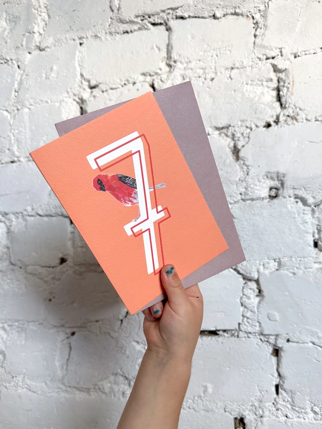bright orange card with a big white number 7 and an illustrated madagascan fody bird with a purple envelope