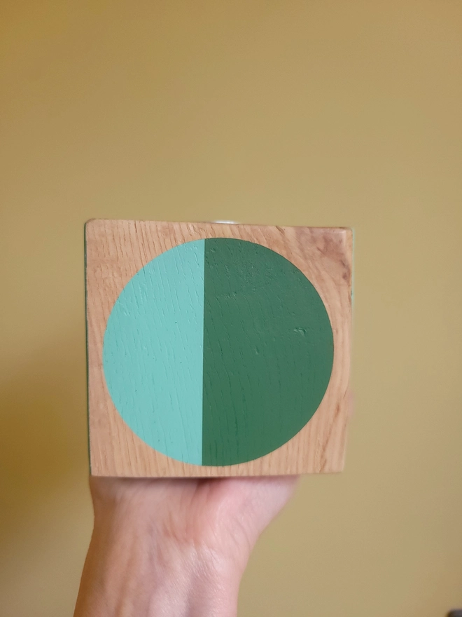 Wood cube tealight holder painted with green and mint circle held up against a yellow wall