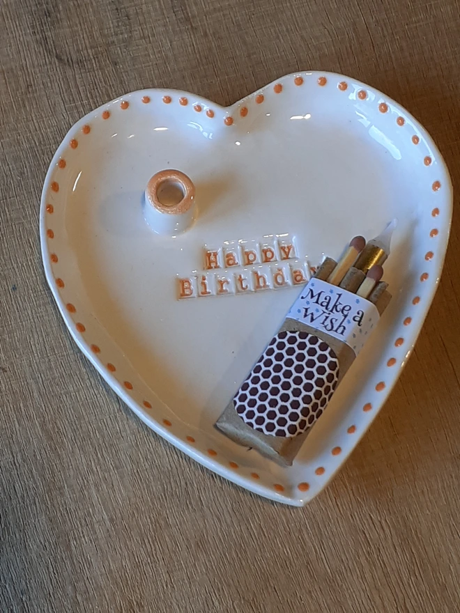 Handmade white heart-shaped ceramic Happy Birthday dish with orange lettering and dotted edging, displayed with a “Make a Wish” matchbox.