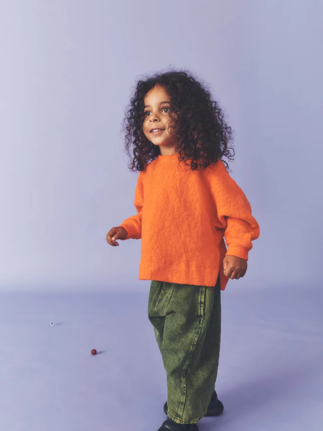 Child mid-step smiling against a lilac backdrop, wearing a Studio Koter orange textured fleece top and green acid-wash barrel-leg jeans