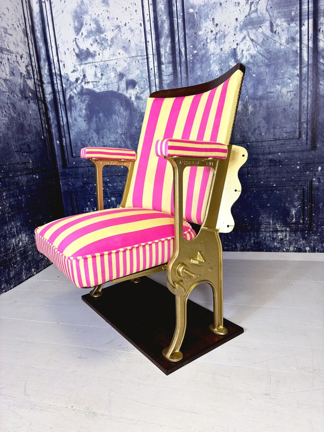 A pink and yellow vintage cinema seat.  The cinema seat has gold legs and is mounted onto a wooden plinth.  The fabric is pink and yellow striped.
