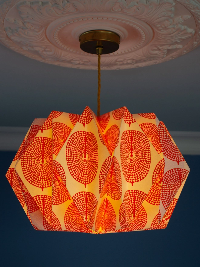 A vintage-inspired paper lampshade with a bold geometric pattern adds a touch of retro flair to a room with a blue wall and ornate ceiling. The lampshade is made from folded paper, creating a unique and eye-catching design.