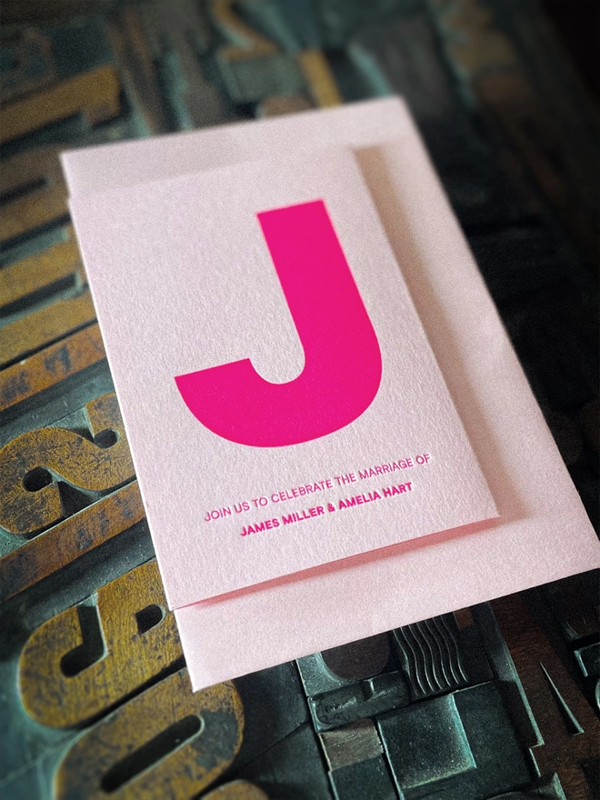Personalised letterpress wedding invitation and matching Candy Pink envelope, featuring oversized fluorescent pink initial and contemporary typographic design.
