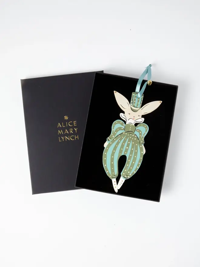Verdant Hare Tree Dancer. the verdant hare tree dancer hanging ornament sits inside its black presentation box, with the lid open to reveal "alice mary lynch" embossed in gold lettering. the hare figure in a striped green and teal jumpsuit with a top hat and bow tie hangs by its teal ribbon and gold bell against the black velvet-lined interior. the branded box gives the piece a polished, gift-ready presentation.