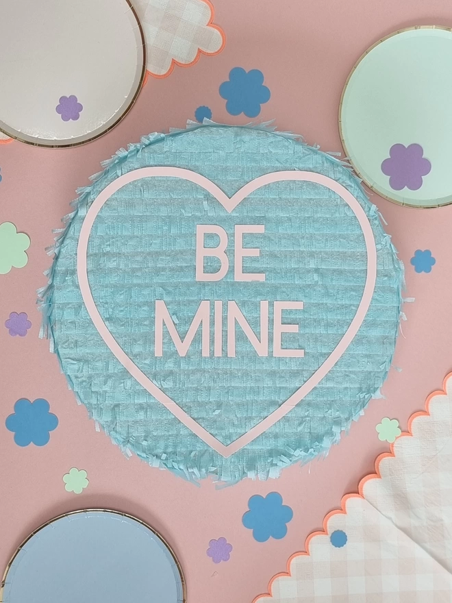 mint green love heart pinata with the words be mine on a pink background surrounded by paper plates and confetti