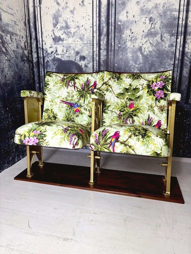 set of two vintage cinema seats upholstered in a purple and green macaws velvet.  Seats are sat on a white wooden floor with a marbled blue wall behind them