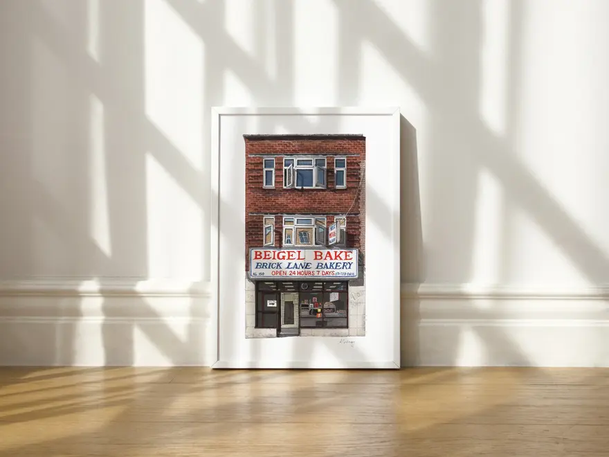 Brick Lane Beigel Bake Giclée Print (Unframed) Shoreditch, a print of a cafe sitting in a white frame on a wooden surface against a plain backdrop. 