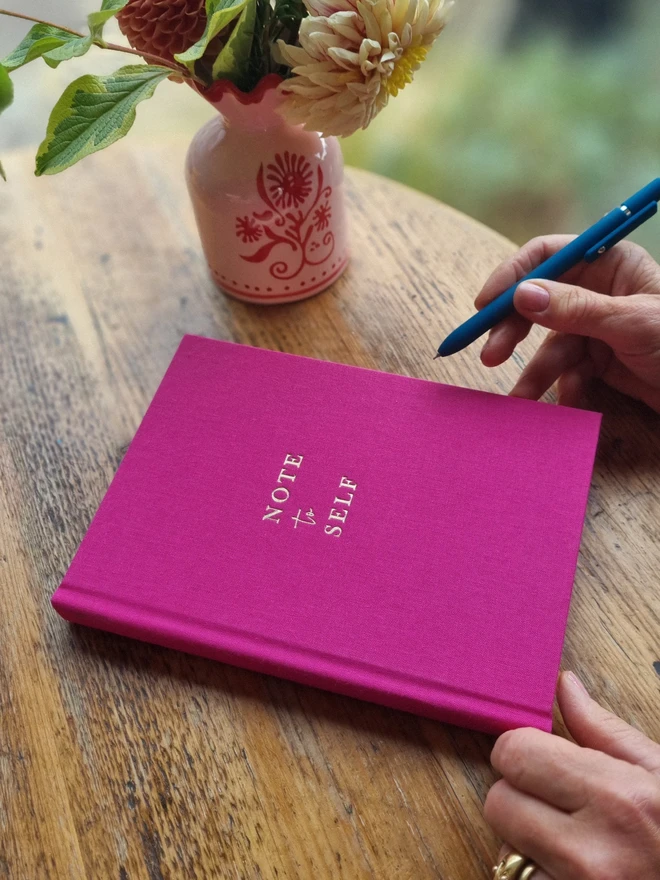 A bright pink notebook printed with the words 'Note to Self' in gold lettering rests on a wooden table top. A woman's hand is shown holding a pen, about to write in the book.