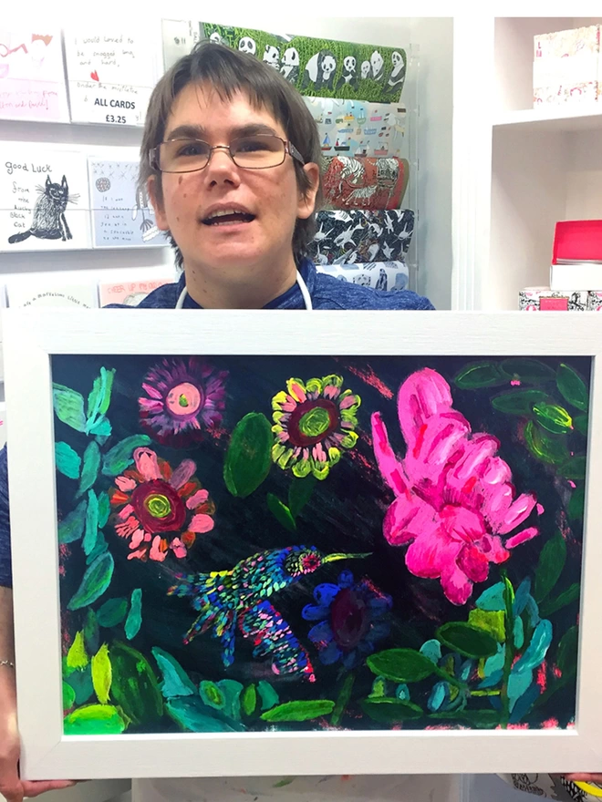 Happy artist holding framed original of a bright coloured painted humming bird for charity chocolate