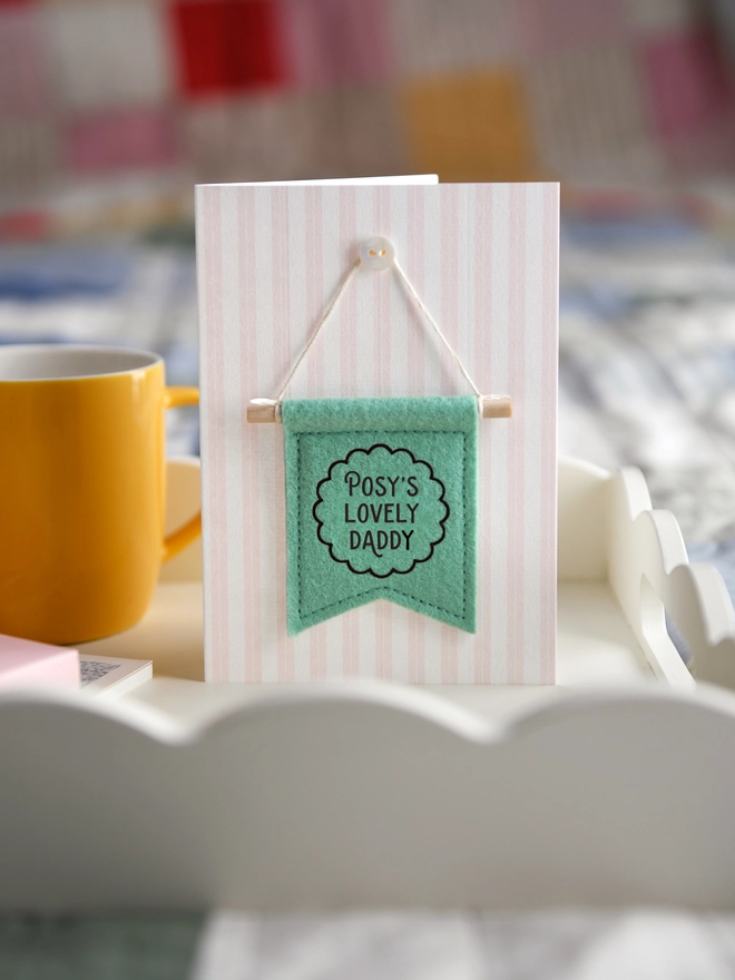 Handmade Father’s Day card with personalised green felt banner “Ivy's Lovely Daddy”, miniature hanging flag keepsake on a wooden breakfast tray. The flag is hung on the card from a tiny mother of pearl button. Beside the card is a book, a mug, a wrapped gift.