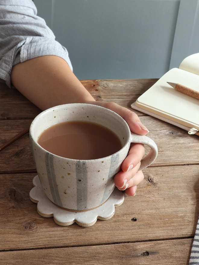Hand holding the handmade Ceramic Stoneware Mug in Blue and White Stripe. The mug sits on top of a flower shaped handmade coaster on top of a wooden surface. A notebook and pen sit behind the mug. 