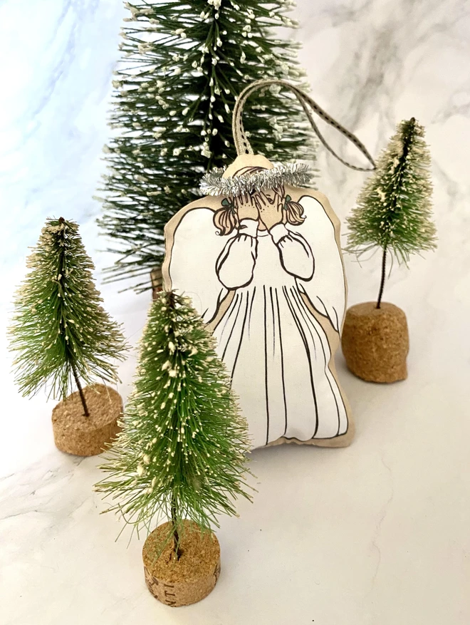 a little christmas tree angel decoration hiding between pint trees playing hide and seek with a little tinsel halo and white dress and bunches made from printed fabric with a ribbon to hang on tree