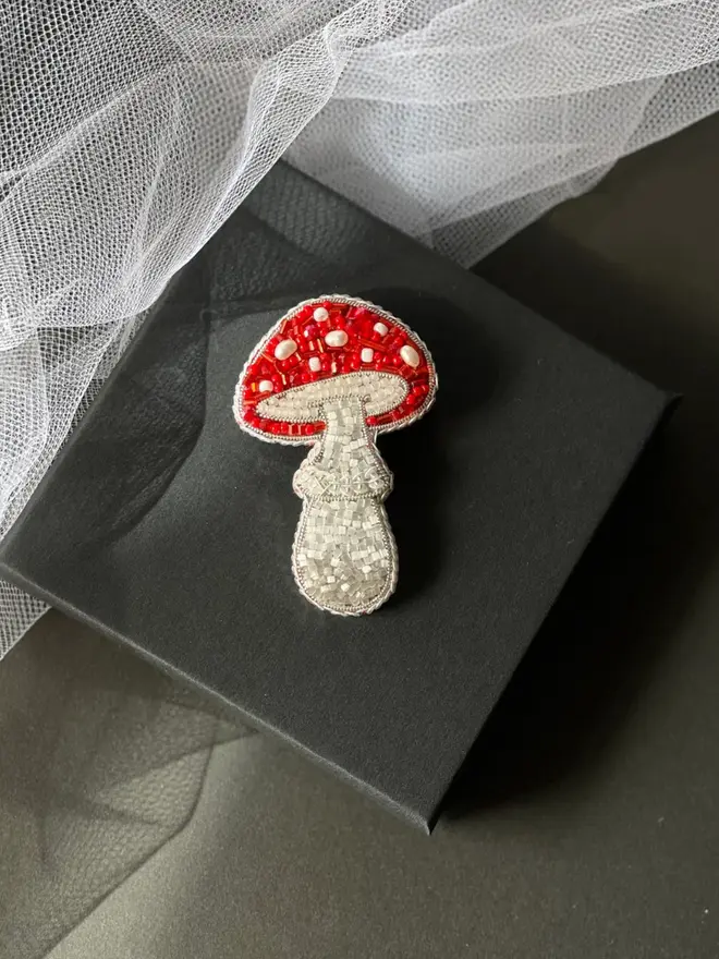 Mushroom Beaded Brooch displayed on a grey box, next to some white fabric. 