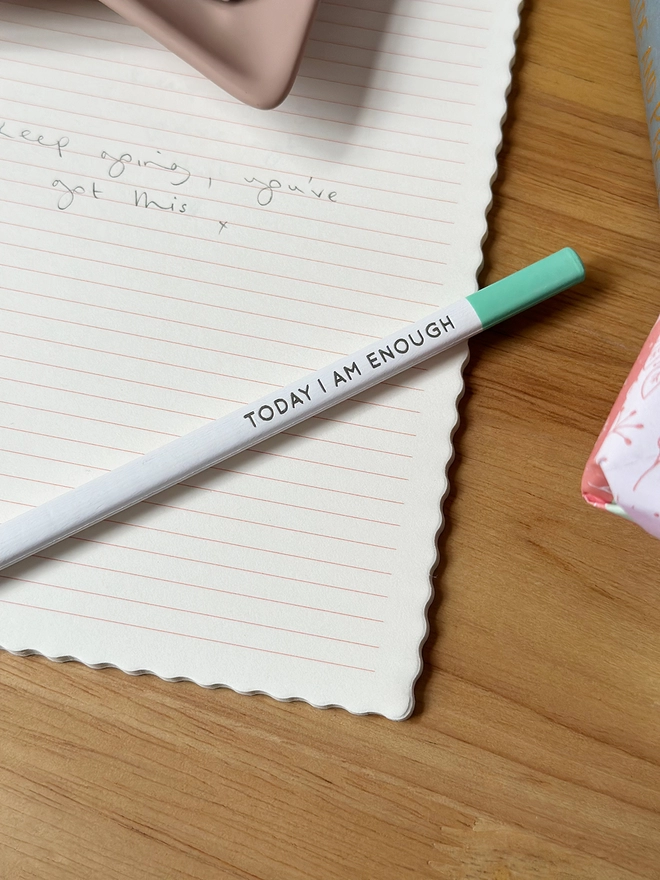 A white pencil, with a green end, and the words "Today I Am Enough" along the side lays on an open lined notebook on a wooden desk.