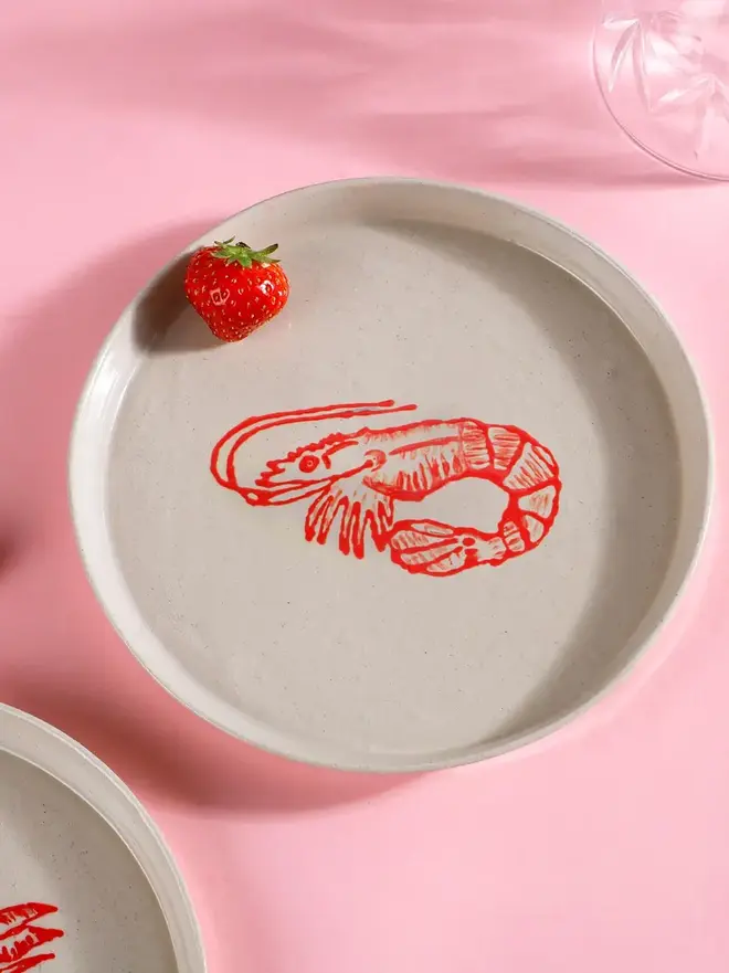 Handmade Ceramic Side Plate With A Red Prawn Design, a plate with a red prawn design sitting on a pink table surface. 