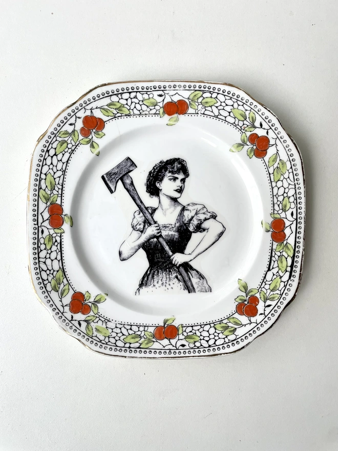 a vintage plate with a really ornate pretty border has a vintage black and white illustration of a beautiful girl in peasant clothes wielding an axe
