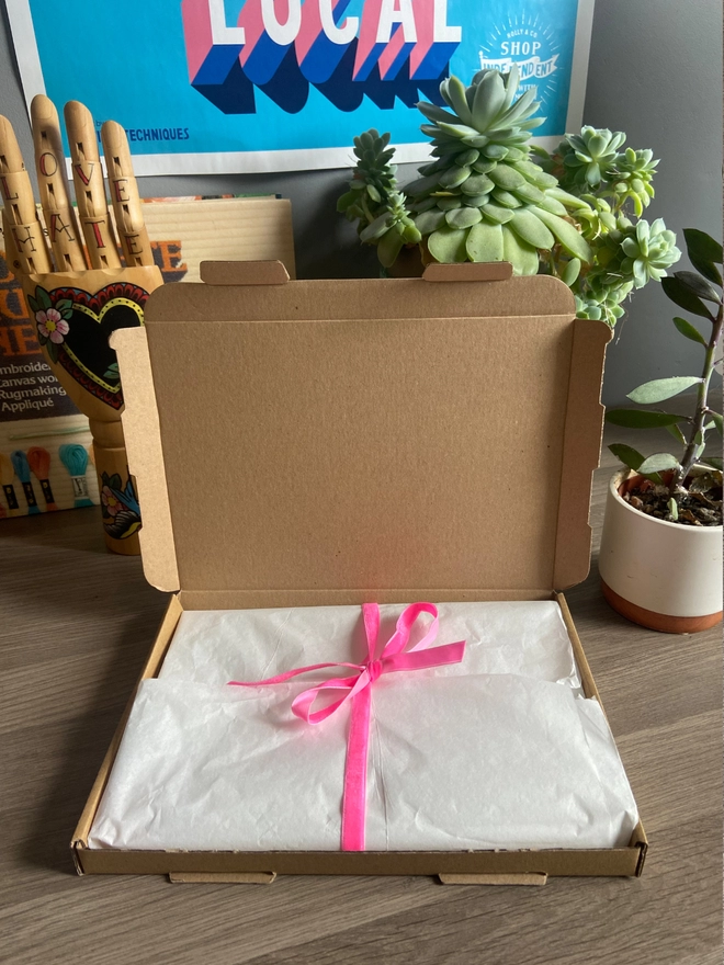 An open box containing an embroidery kit wrapped in tissue paper and tied with pink ribbon 