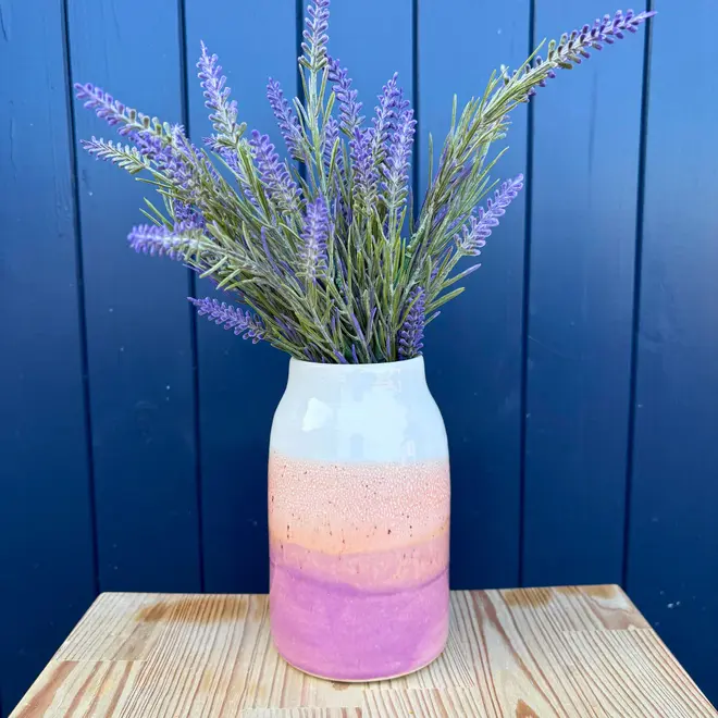Ice Cream Sundae Large Vase, a colourful vase sitting on a wooden surface against a navy blue backdrop. It is accompanied by a plant. 