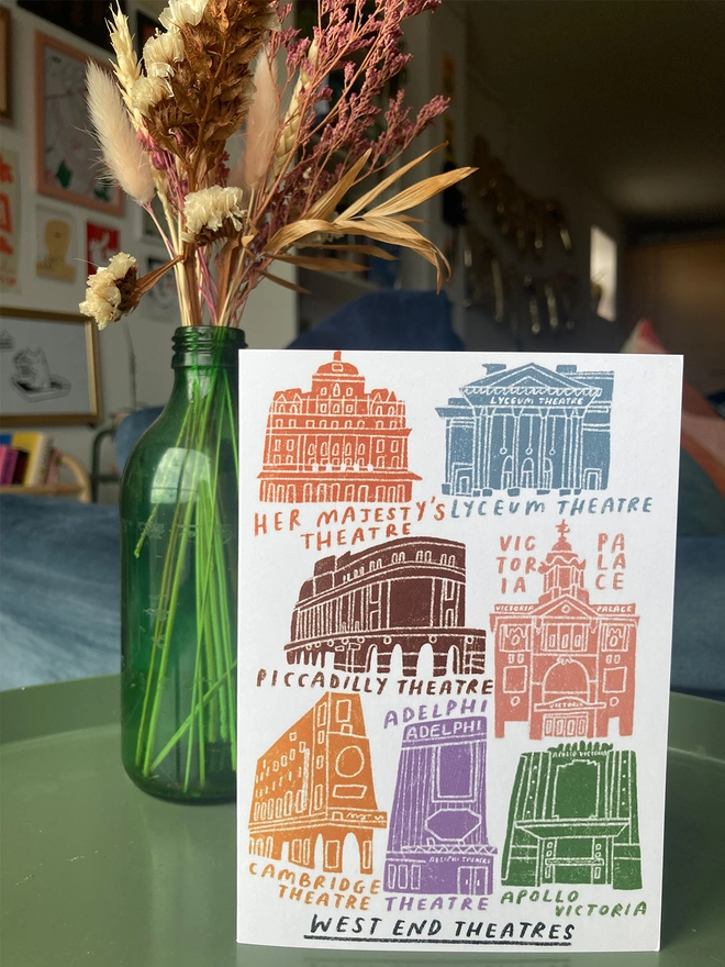 Greetings card with illustrations of West End theatres, placed on a table with flowers in the background