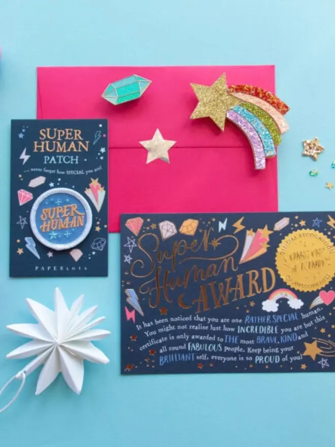 A navy "Super Human Award" certificate with gold foil lettering and an "Official Recognition One of a Kind" seal. The patch card and a pink envelope sit beside it, decorated with a glitter shooting star.