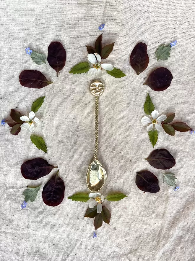 An image of a gold coloured brass spoon featuring a twisted handle and a hand carved moon fave at the top. The spoon is pictured laying on raw linen, surrounded with a symmetrical arrangement of leaves and flowers 